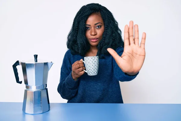 Beautiful african woman drinking a coffee from italian coffee maker with open hand doing stop sign with serious and confident expression, defense gesture