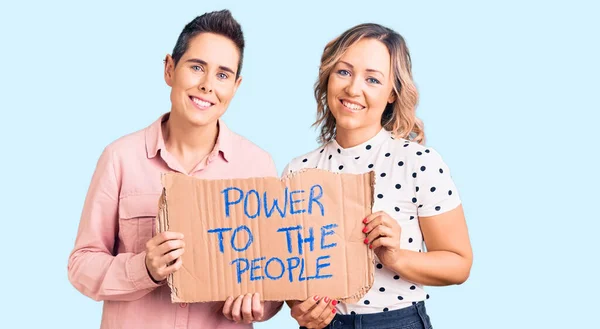 Couple of women holding power to the people banner looking positive and happy standing and smiling with a confident smile showing teeth