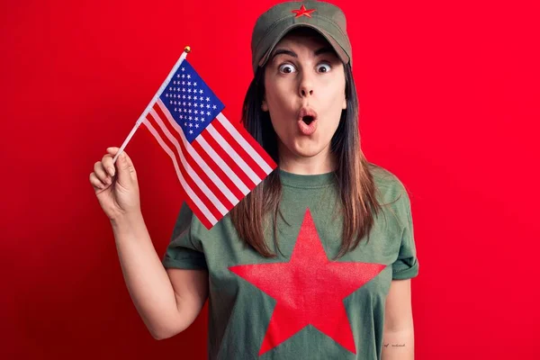 Beautiful woman wearing t-shirt with red star communist symbol holding united states flag scared and amazed with open mouth for surprise, disbelief face