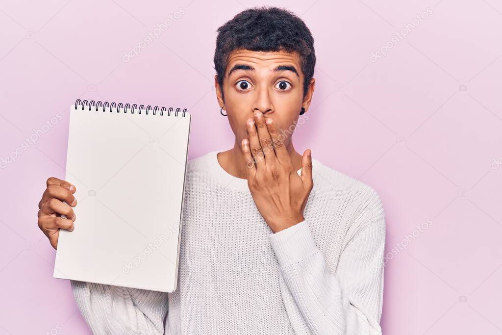 Young african amercian man holding notebook covering mouth with hand, shocked and afraid for mistake. surprised expression 
