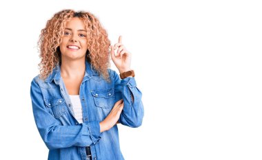 Kıvırcık saçlı, sıradan kot ceket giyen, yüzünde kocaman bir gülümseme olan, eli ve parmağı kameraya bakan genç sarışın kadın.. 