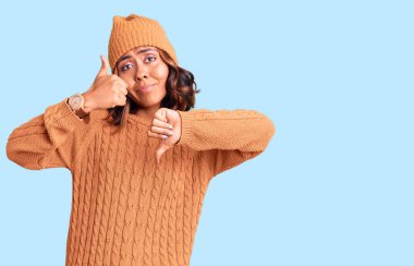 Genç ve güzel melez kadın yün kazak ve kış şapkası giyip baş parmak kaldırıyor, anlaşmazlık ve anlaşma ifadesi takınıyor. çılgın çatışma 