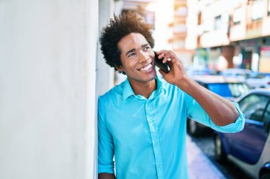 Gülümseyen mutlu gülümseyen Afrikalı Amerikalı genç bir adam. Yüzünde gülümsemeyle duvara yaslanarak şehrin caddesinde akıllı telefondan konuşarak..