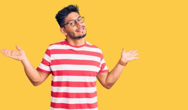 Yakışıklı Latin Amerikalı genç adam gündelik kıyafetler ve gözlük takıyor. Hiçbir şeyden haberi yok ve elleri ve kolları havada şaşkın bir ifade takınıyor. şüphe kavramı. 