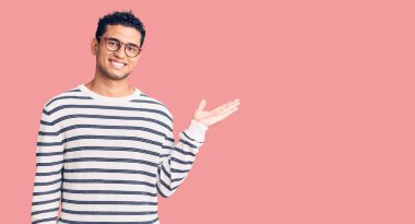 İspanyol asıllı yakışıklı genç adam gündelik kıyafetler ve gözlük takıyor neşeli bir şekilde gülümsüyor ve avuç içi kameraya bakıyor.. 