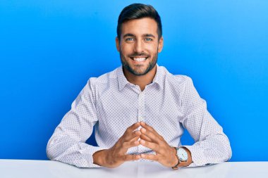 İş kıyafetleri giymiş yakışıklı İspanyol adam masanın üzerinde el ele oturuyor ve mutlu ve rahat bir şekilde gülümsemeyi diliyordu. Başarı ve iyimserlik 