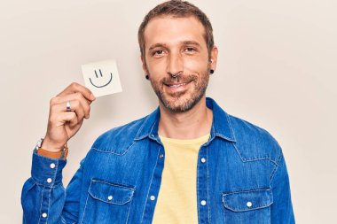 Elinde gülümseme emojisi olan genç bir adam pozitif ve mutlu görünüyor. Kendine güvenen bir gülümsemeyle ayakta duruyor ve dişlerini gösteriyor. 