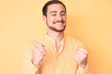 Gündelik kıyafetler giyen, mutlu ve heyecanlı genç bir adam kollarını kaldırmış, gülümseyen ve başarı için çığlık atan kazanan bir jest yapıyor. kutlama konsepti. 