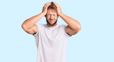 Beyaz gündelik tişört giyen genç beyaz bir adam baş ağrısı çekiyor. Acı ve migren yüzünden çaresiz ve stresli. Eller başının üstünde.. 