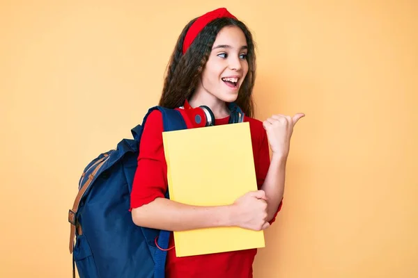 Linda Niña Hispana Usando Mochila Estudiante Auriculares Sosteniendo Libro Apuntando —  Fotos de Stock