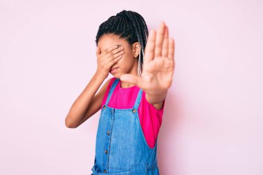 Saçları örgülü, pembe arka plan üzerine günlük kıyafetler giyen, gözleri ellerle kaplayan, üzgün ve korku ifadeli hareketlerini durduran Afro-Amerikalı genç kız. utangaç ve olumsuz kavram. 