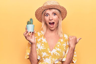 Sarı arka planda elinde kaktüs saksısı tutan güzel sarışın kadın baş parmağını yukarı kaldırıyor ve ağzı açık gülümsüyor.