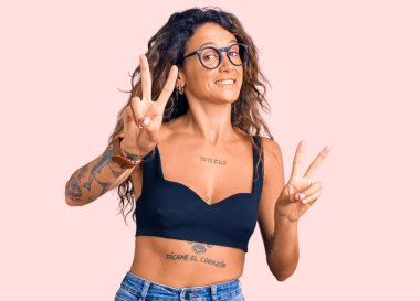 Günlük kıyafetler ve gözlük takan, gülümseyen, parmaklarını zafer işareti yaparken gösteren Latin kökenli genç bir kadın. İki numara.. 