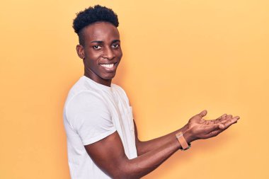 Afro-Amerikan genç adam, günlük giysiler içinde avuçları açık, fotokopi alanını gösteren, gülümseyerek mutlu bir gülümseme sunan. 