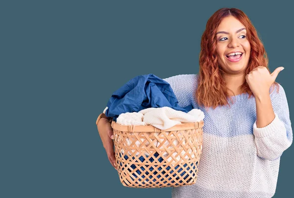 Young Latin Woman Holding Laundry Basket Pointing Thumb Side Smiling — Stock Photo, Image