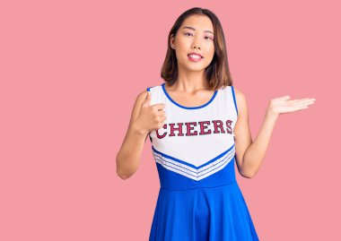 Ponpon kız üniforması giyen genç güzel Çinli kız avuç içini gösteriyor ve baş parmaklarıyla iyi bir jest yapıyor, mutlu ve neşeli gülümsüyor. 