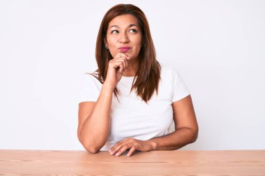 Orta yaşlı esmer, Latin bir kadın, gündelik beyaz tişört giyip masaya oturmuş, parmakları çenesinde şüpheye odaklanmış ve kafasını kaldırıp, 