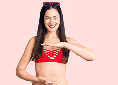 Genç, beyaz, güzel bir kadın. Bikini giyiyor. Ellerinde kocaman bir işaret, ölçü işareti var. Kameraya bakıp gülümsüyor. ölçüm kavramı. 