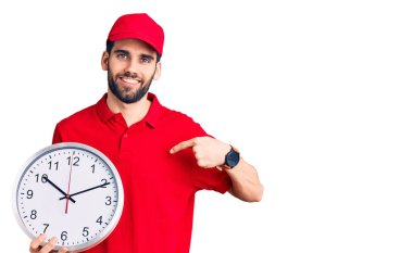 Sakallı, teslimat üniformalı, koca saati tutan yakışıklı genç adam gülümseyen, mutlu ve gururlu birini işaret ediyor. 