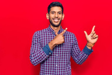 Gündelik kıyafetler giyen, gülümseyen ve kameraya bakan genç latin adam iki eliyle ve parmaklarıyla kenarı işaret ediyor.. 