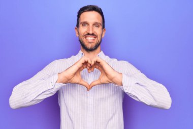 İzole edilmiş mor arka planda dikilmiş, sıradan çizgili tişört giyen genç yakışıklı bir adam elleriyle kalp sembolü yapan bir aşkla gülümsüyor. Romantik konsept.