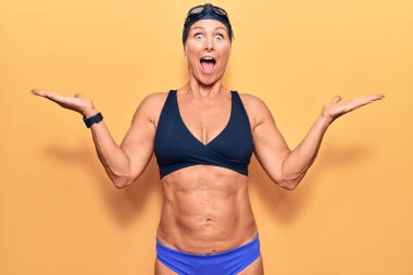 Orta yaşlı sarışın kadın bikini giyiyor, yüzme şapkası takıyor ve Google 'da zaferi kutluyor mutlu bir gülümsemeyle ve ellerini kaldırarak kazanan bir ifadeyle. 