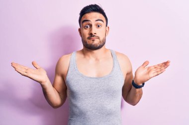 Sakallı, pembe arka planda kolsuz tişört giyen, kolları açık ve kafası karışmış genç yakışıklı bir adam..