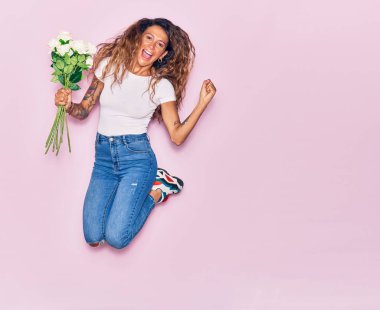 Genç, güzel, kıvırcık bir kadın elinde bir buket beyaz gül ile mutlu bir şekilde gülümsüyor. Yüzünde tebessümle zıplıyor, pembe arka planda yumruklarıyla kutluyor.