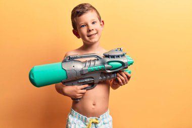 Mayo giyen tatlı sarışın çocuk elinde su tabancası tutuyor. Pozitif görünüyor ve mutlu görünüyor. Kendine güvenen bir gülümsemeyle dişlerini gösteriyor. 