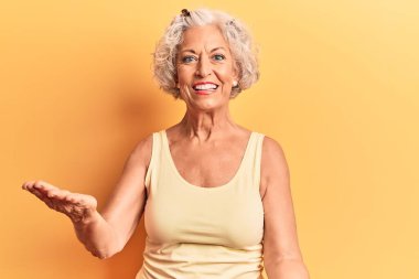 Kıdemli, gri saçlı, günlük kıyafetler giyen neşeli, kollarını açıp gülümseyen bir kadın. Hoş geldin, olumlu ve kendinden emin selamlar. 