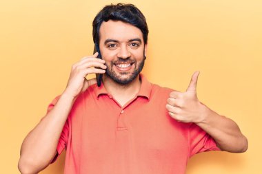 Genç İspanyol adam akıllı telefondan konuşuyor mutlu ve olumlu gülümsüyor, başparmağıyla mükemmel ve onay işareti yapıyor. 