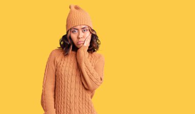 Genç, güzel, melez, yün kazak giyen, kışlık şapkalı, yorgun görünen ve depresyondan sıkılmış kolları çaprazlanmış bir kadın.. 