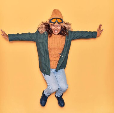 Kışlık giysiler giyen genç, güzel Latin kadın mutlu gülümsüyor. Yüzünde ve kollarında tebessümle, izole edilmiş sarı arka planda atlıyor.