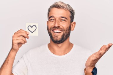 Sarışın ve sakallı yakışıklı bir adam. Beyaz arka planda kalp şeklinde hatırlatıcı taşıyor. Başarıyı mutlu bir gülümsemeyle kutluyor.