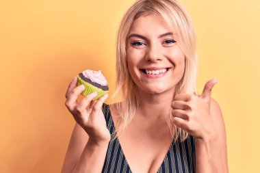 Sarışın, güzel bir kadın, izole edilmiş sarı arka planda tatlı çikolatalı kek yiyor mutlu ve pozitif gülümsüyor, başparmağını kaldırıyor ve mükemmel bir şekilde işaret veriyor.