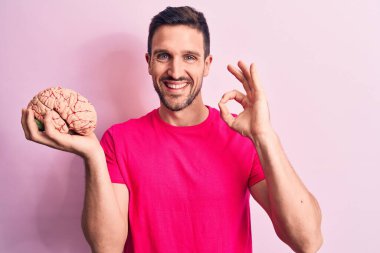 Genç ve yakışıklı bir adam beyni soyutlanmış pembe arka planda tutarak hafızasını tazelemek istiyor. Parmaklarıyla işaret ediyor, gülümseyerek mükemmel bir işaret veriyor.