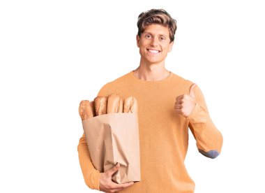 Elinde ekmek dolu kağıt torba tutan genç ve yakışıklı adam mutlu ve olumlu gülümsüyor, başparmağı mükemmel ve onay işareti yapıyor. 
