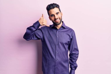 Sakallı, sıradan tişörtlü, gülümseyen, el ve parmaklarıyla telefon hareketi yapan, telefonda konuşan genç, yakışıklı bir adam. iletişim kavramları. 