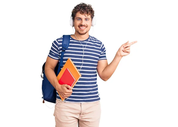 年轻英俊的男人 卷曲的头发 提着学生的背包和书本 笑容满面 手指手画脚地指向旁边 — 图库照片