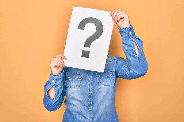 Woman covering face holding paper with question mark symbol. Standing over isolated yellow background