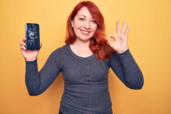 Young beautiful redhead woman holding broken smartphone showing cracked screen doing ok sign with fingers, smiling friendly gesturing excellent symbol