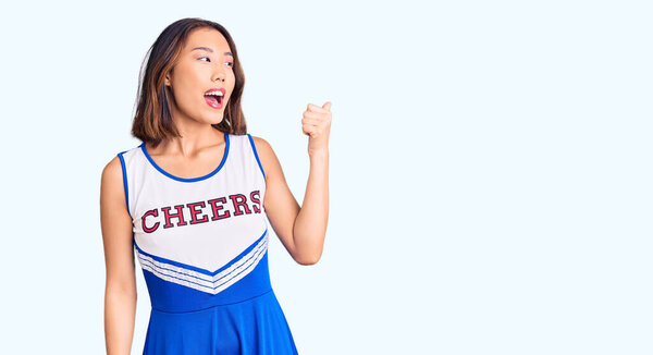 Young beautiful chinese girl wearing cheerleader uniform smiling with happy face looking and pointing to the side with thumb up. 