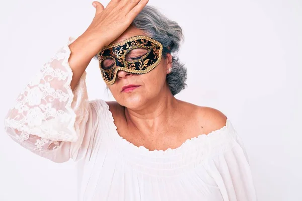 Senior hispanic grey- haired woman wearing venetian carnival mask surprised with hand on head for mistake, remember error. forgot, bad memory concept.
