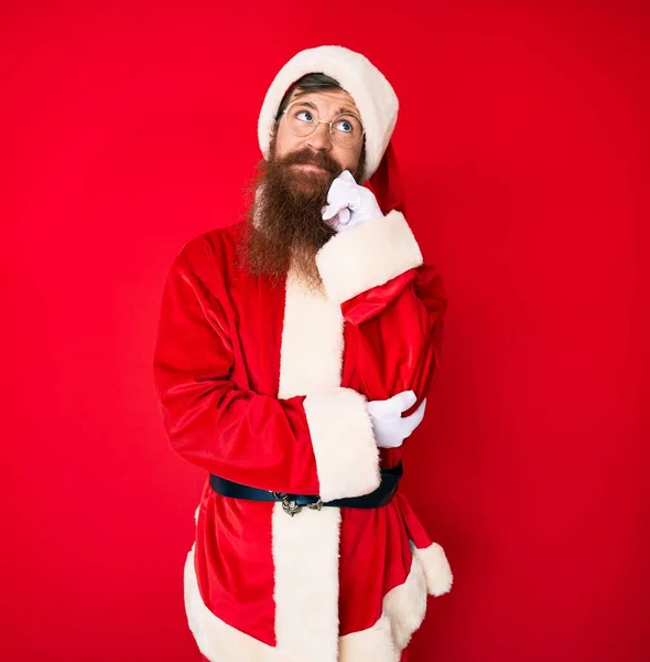 Handsome young red head man with long beard wearing santa claus costume serious face thinking about question with hand on chin, thoughtful about confusing idea