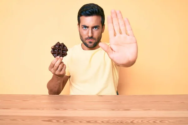 Jovem Hispânico Segurando Waffle Chocolate Sentado Mesa Com Mão Aberta — Fotografia de Stock