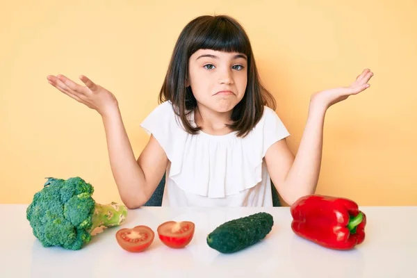 若いです女の子ともに座っているテーブルの上に野菜無謀と混乱していますオープン腕 見当もつかないと疑わしい顔 — ストック写真