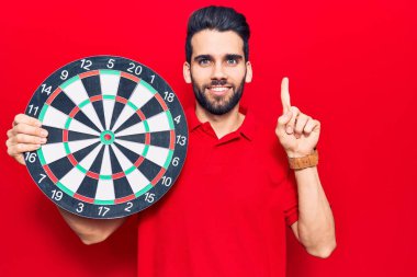 Sakallı, elinde dart tahtası olan genç yakışıklı bir adam bir fikirle gülümsüyor ya da bir soru işareti yapıyor. 