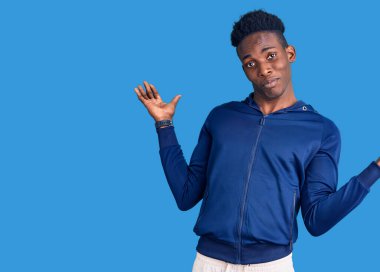 Genç Afrikalı Amerikalı adam spor kıyafetleri giyiyor ve elleri ve kolları havada kafası karışmış bir ifade takınıyor. şüphe kavramı. 