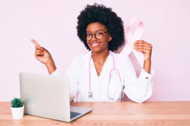 Genç Afro-Amerikan kadın doktor üniforması giyiyor elinde pembe kanser kurdelesi, gülümsüyor. 