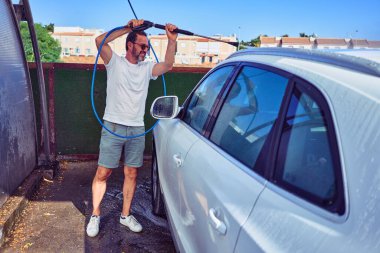 Orta yaşlı yakışıklı, günlük kıyafetler giyen ve mutlu gülümseyen güneş gözlüklü bir adam. Yüzünde gülümsemeyle ayakta durup su tabancası kullanarak araba yıkamak..